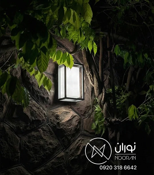 چراغ دیواری بیرونی مدرن مشکی مدل H214 با پنل نوری سفید مستطیلی، نصب شده روی دیوار سنگی حیاط در میان گیاهان با نور آفتابی.