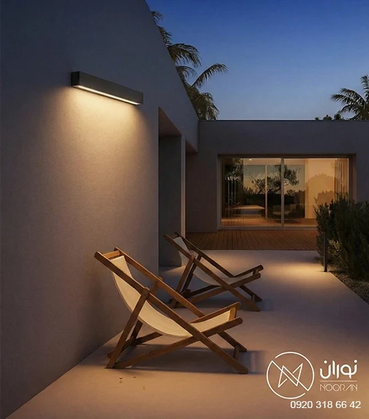چراغ دیواری بیرونی مدرن و خطی خاکستری مدل H131-1 با پخش نور یک‌طرفه رو به پایین، نصب شده روی دیوار ویلا بالای صندلی‌های استراحت در شب.