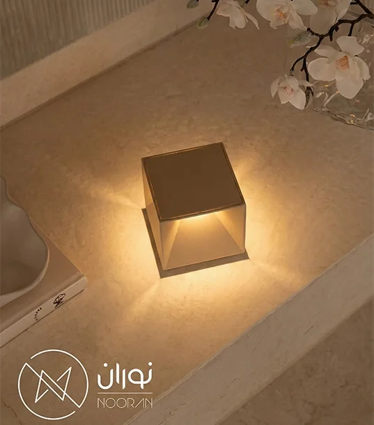 چراغ رومیزی شارژی مدرن مدل Cubox با طراحی مکعبی مینیمال به رنگ برنز، در حال تابش نور گرم روی میز سنگی در کنار کتاب و گل‌های سفید.